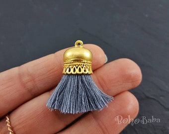 May include: A gray tassel with a gold metal top. The top has a decorative design and a loop for hanging.
