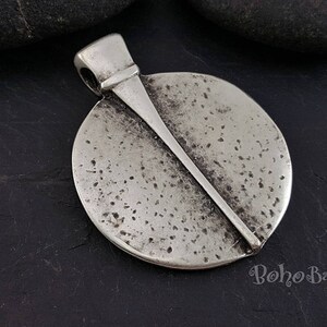 May include: A silver-toned pendant with a circular design and a diagonal bar across the face. The pendant has a textured surface with small dark spots and a loop for a chain. The word "Boho Baba" is visible in the bottom right corner.
