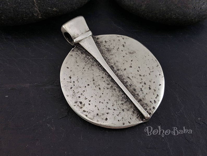 May include: A silver-toned pendant with a circular design and a diagonal bar across the face. The pendant has a textured surface with small dark spots and a loop for a chain. The word "Boho Baba" is visible in the bottom right corner.