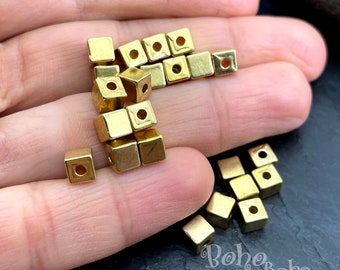 May include: A pile of gold-colored metal square beads with a hole in the center of each bead. The beads are arranged in a random pattern on a hand.