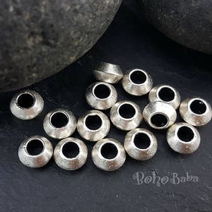 May include: A close-up of a group of silver metal beads with a round shape and a hole in the center. The beads are arranged in a random pattern on a dark gray surface.