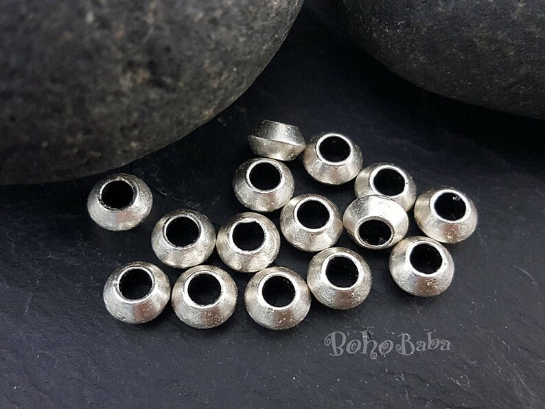 May include: A close-up of a group of silver metal beads with a round shape and a hole in the center. The beads are arranged in a random pattern on a dark gray surface.
