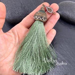 May include: A sage green tassel pendant featuring an ornate bronze-toned metal cap with a Greek key pattern, small clear rhinestones, and green accents. A small leaf-shaped charm with clear rhinestones is attached to the top loop of the tassel.