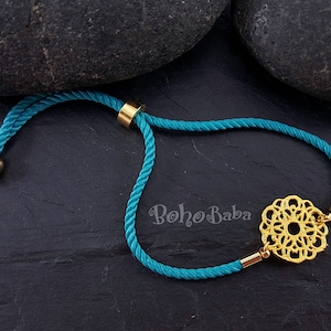 May include: Teal blue adjustable bracelet with a gold filigree charm and gold beads. The bracelet is tied with a slip knot and has the text "boho baba" printed on the background.