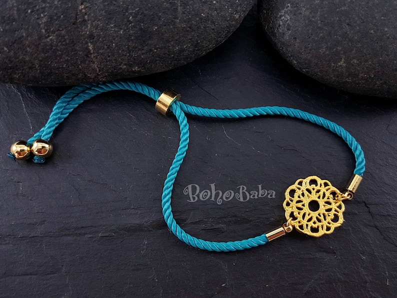 May include: Teal blue adjustable bracelet with a gold filigree charm and gold beads. The bracelet is tied with a slip knot and has the text "boho baba" printed on the background.