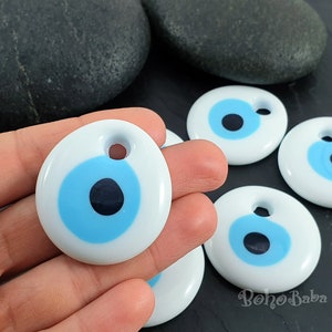May include: Five white ceramic evil eye charms with blue and black eyes. The charms have a hole at the top for hanging.