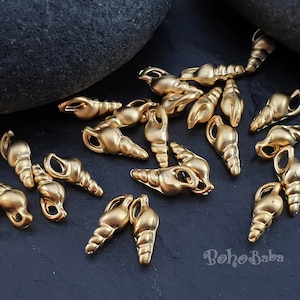 Puede incluir: Una colección de abalorios de concha de mar de color dorado con un bucle en la parte superior. Los abalorios están esparcidos sobre una superficie gris oscura.