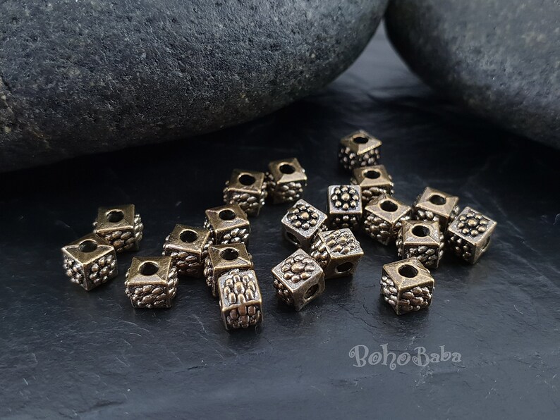 May include: A group of 20 antique gold square beads with a decorative design. The beads have a hole in the center for stringing.