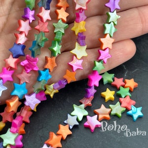 Op de afbeelding: Strengen kleurrijke, stervormige kralen in tinten roze, oranje, groen, blauw en geel. De kralen zijn geregen, met enkele losse kralen verspreid in de buurt. De kralen hebben een parelmoerglans.