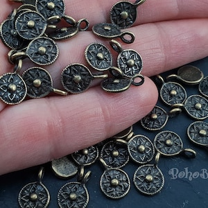 May include: A close-up of a pile of antique bronze metal charms with a starburst design and a loop for attaching to jewelry or other crafts. The charms are arranged on a hand.