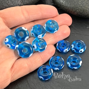 Op de afbeelding: Een verzameling doorschijnende blauwe glazen kralen met witte en zwarte oogontwerpen. De ronde kralen hebben een gat in het midden. De kralen zijn helderblauw en worden in een hand gehouden.