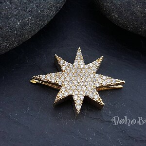 Puede incluir: Encanto de estrella dorado con diamantes de imitaci&oacute;n transparentes. La estrella tiene ocho puntas y est&aacute; sobre un fondo gris oscuro.