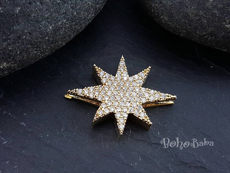 Puede incluir: Encanto de estrella dorado con diamantes de imitaci&oacute;n transparentes. La estrella tiene ocho puntas y est&aacute; sobre un fondo gris oscuro.