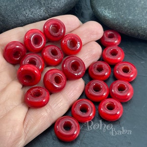 Puede incluir: Una colección de cuentas rojas brillantes en forma de rosquilla. Las cuentas tienen un agujero en el centro y una superficie lisa y pulida. Las cuentas están esparcidas sobre una superficie oscura. Las cuentas son de color rojo vibrante.