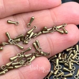 May include: A close-up of a pile of antique gold-toned metal teardrop-shaped pendant charms with a loop at the top. The charms are scattered on a hand.