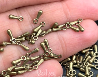 May include: A close-up of a pile of antique gold-toned metal teardrop-shaped pendant charms with a loop at the top. The charms are scattered on a hand.