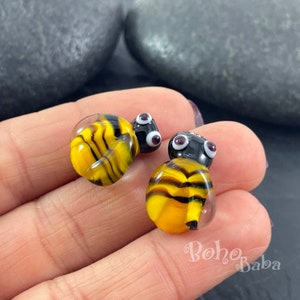 Puede incluir: Dos pequeñas cuentas de vidrio en forma de abeja con rayas negras y amarillas y ojos blancos. Las cuentas miden aproximadamente 1 cm de diámetro.