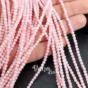 Può includere: Primo piano di fili di piccole perline rotonde rosa chiaro. Le perline sono infilate insieme, creando più linee verticali. Le perline hanno un aspetto liscio e lucido. Lo sfondo è scuro, il che fa risaltare le perline rosa. L'immagine ha un aspetto morbido e delicato.