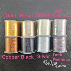 Puede incluir: Ocho carretes de hilo en varios colores, incluyendo dorado, beige, crema, arcoíris claro, cobre, negro, plateado y arcoíris oscuro. Los carretes están etiquetados con sus respectivos colores. La marca "Boho Baba" es visible en la esquina inferior derecha.