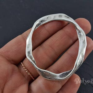 Puede incluir: Un anillo de círculo abierto de tono plateado con una forma irregular texturizada. El anillo está hecho de metal y tiene un acabado liso y pulido.