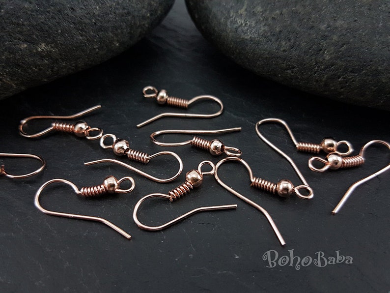 May include: Rose gold earring hooks with a spring and a small ball at the top. The hooks are scattered on a dark grey surface.