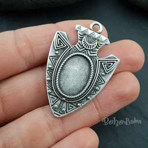 Può includere: Ciondolo a forma di punta di freccia color argento con incastonatura ovale e intricati motivi geometrici. Il ciondolo ha un anello nella parte superiore per il fissaggio a una catena o un cordino. Il design include triangoli, zig-zag e una forma a mezzaluna, creando un'estetica tribale o bohémien.