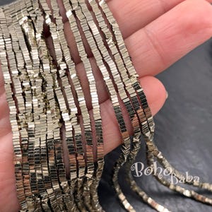Op de afbeelding: Close-up van een streng metallic, rechthoekige kralen in zilverkleur. De kralen worden in een hand gehouden, met de merknaam "Boho Baba" zichtbaar in de rechter benedenhoek. De kralen zijn waarschijnlijk voor het maken van sieraden.