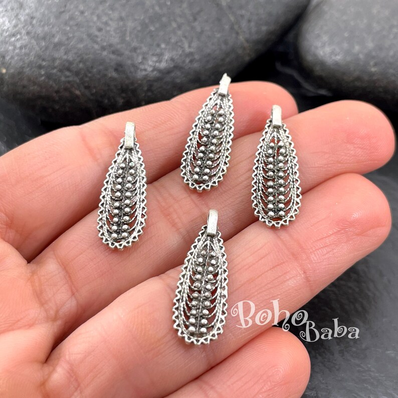 May include: Four silver-colored, teardrop-shaped pendants with intricate openwork and a beaded center design. Each pendant has a small loop at the top for attaching to a chain or earring hook. The pendants are displayed on a hand.