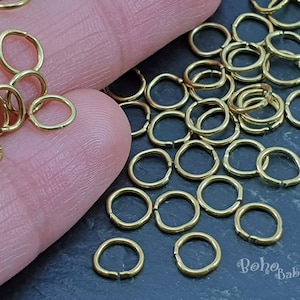 May include: A large pile of gold jump rings, also known as split rings, are scattered on a dark gray surface. The rings are small and perfect for jewelry making.