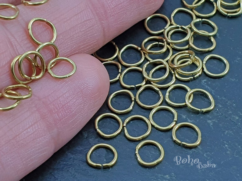 May include: A large pile of gold jump rings, also known as split rings, are scattered on a dark gray surface. The rings are small and perfect for jewelry making.