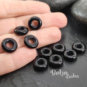 Puede incluir: Diez cuentas negras en forma de rosquilla con un agujero en el centro. Las cuentas están dispuestas en semicírculo sobre una mano.