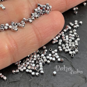 Puede incluir: Primer plano de un montón de cuentas de metal plateado. Las cuentas son pequeñas y de forma cuadrada. Las cuentas están esparcidas sobre una superficie oscura.