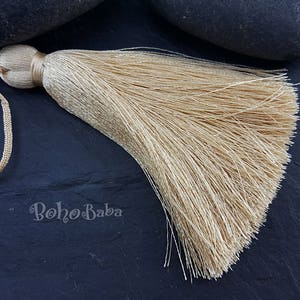 May include: A beige tassel with a long, flowing fringe. The tassel is made of soft, silky fibers and has a loop at the top for hanging.