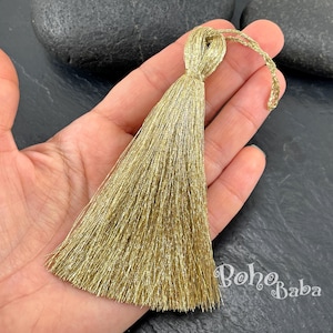 May include: A metallic gold tassel featuring a dense cluster of fine, shimmering threads gathered at the top with a wrapped cord and a small loop for hanging. The tassel has a straight, fan-like shape and a bright, reflective finish throughout.