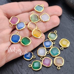 Op de afbeelding: Assortiment van 14 goudkleurige metalen bedels met gekleurde glazen inzetstukken. Elke bedel heeft een vierkante vorm met een lus om te bevestigen aan sieraden of andere knutselwerkjes.