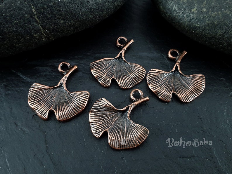 May include: Four copper-colored Ginkgo leaf charms with a textured, veined design. Each charm has a loop at the top for attaching to jewelry. The charms are displayed on a dark, textured surface.