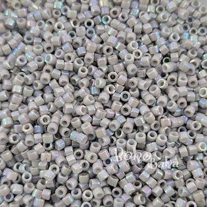 May include: A close-up image of a pile of small, iridescent, light gray seed beads. The beads are round and have a smooth, shiny finish.