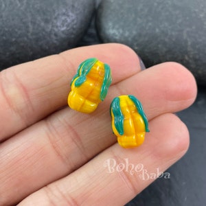 Puede incluir: Dos cuentas de vidrio amarillo con hojas verdes. Las cuentas tienen forma de mazorcas de maíz.