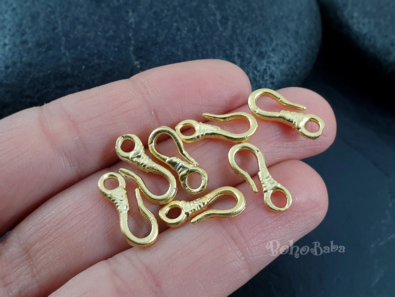 May include: A close-up of ten gold-toned metal lobster claw clasps. The clasps are arranged on a hand, with the tips of the clasps facing the camera.