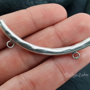 Puede incluir: Conector de metal plateado con forma curva y dos pequeños bucles. El conector tiene una textura martillada.