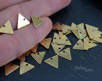 Op de afbeelding: Een stapel gouden metalen driehoekige bedels met een gat in het midden. De bedels liggen verspreid op een zwart oppervlak.