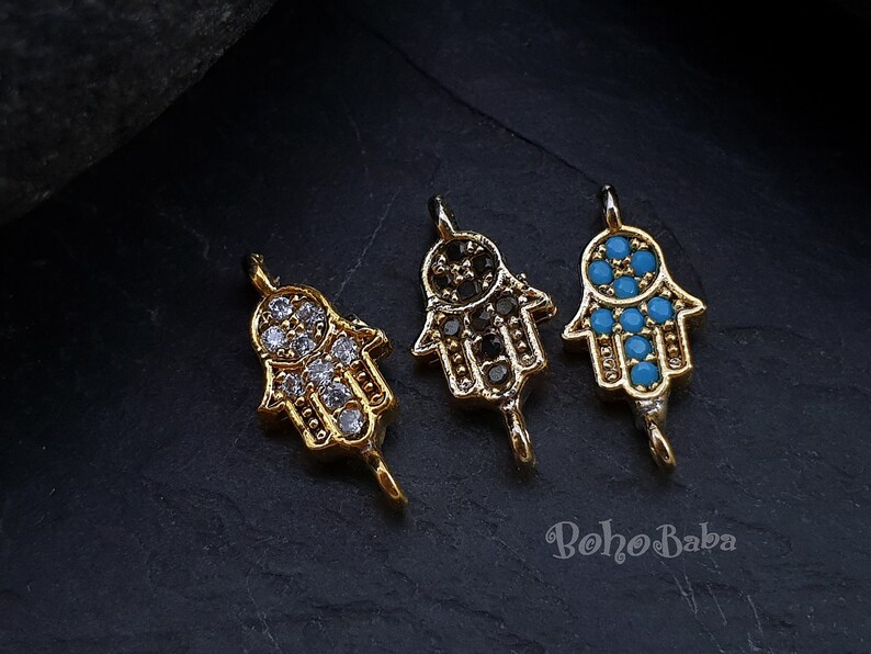 Puede incluir: Tres dijes de mano de Hamsa dorados con piedras de colores diferentes. Los dijes est&aacute;n sobre un fondo negro. La mano de Hamsa es un s&iacute;mbolo de protecci&oacute;n y buena suerte.