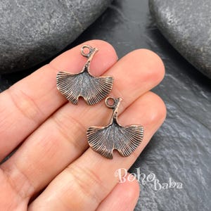 May include: Two bronze-colored ginkgo leaf charms with detailed veining. Each charm has a loop at the top for attaching to jewelry. The charms are displayed on a hand, with the text "Boho Baba" visible in the background.