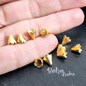 May include: A close-up of ten gold-coloured metal flower-shaped bead caps. The caps are arranged on a hand, with the tips of the caps pointing towards the camera.