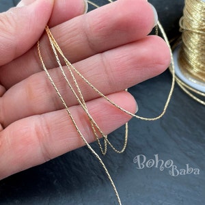Op de afbeelding: Close-up van een gouden metallic koord, een soort touw of draad, gewikkeld om een spoel. Het koord wordt in een hand gehouden, waardoor de textuur en glans zichtbaar zijn.