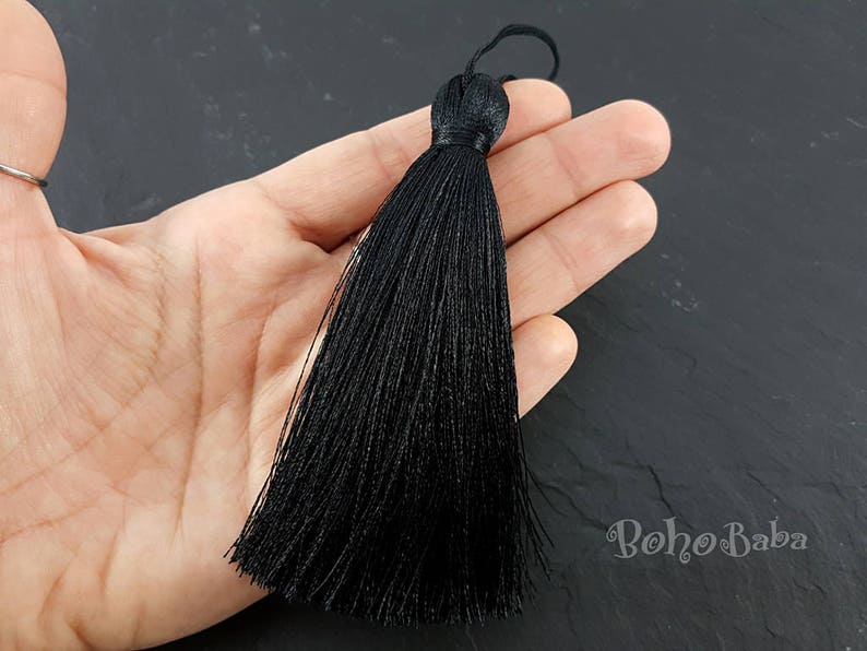 K&ouml;nnte beinhalten: Eine lange, schwarze Quaste mit einer Schlaufe oben. Die Quaste besteht aus vielen Str&auml;ngen schwarzen Fadens, die oben zusammengebunden sind. Die Quaste wird in einer Hand vor einem dunklen Hintergrund gehalten.