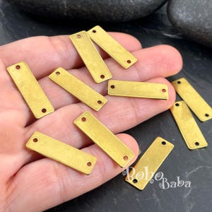 May include: A collection of small, rectangular brass-toned metal tags, each featuring two circular holes at opposite ends. These flat, elongated components have a matte finish and are arranged in a scattered pattern, showing a consistent design.
