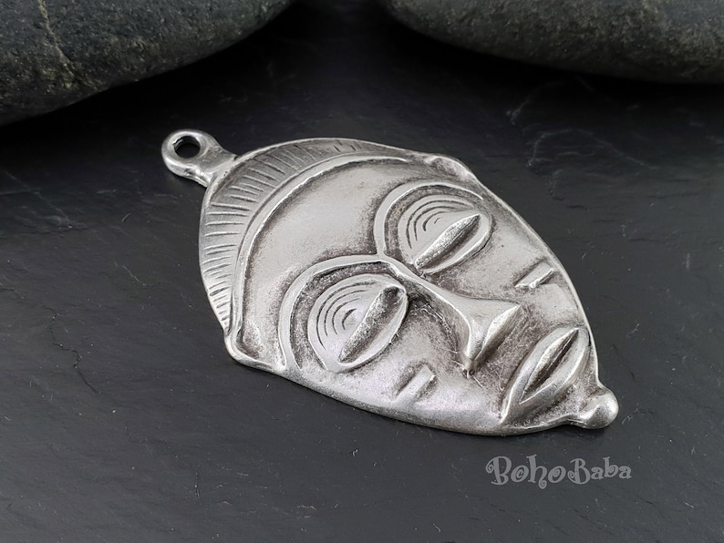 May include: Silver pendant featuring a detailed, stylized African mask with closed eyes and a textured surface.