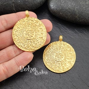 May include: Two gold-toned round pendants with intricate circular designs and raised lettering. The pendants are approximately 2 inches in diameter.
