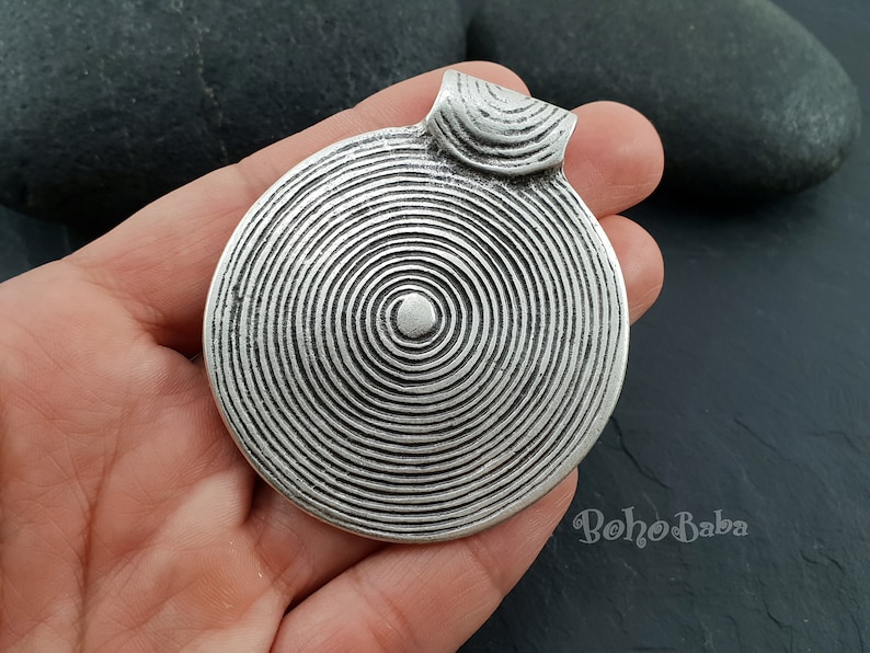 May include: Silver pendant with a circular design featuring concentric circles. The pendant has a loop at the top for attaching a chain.
